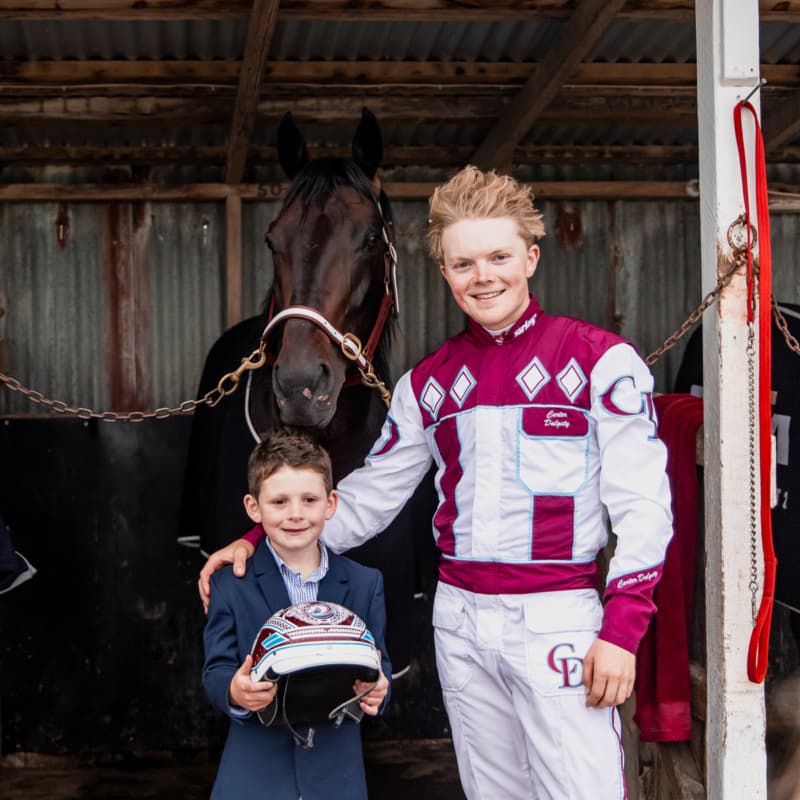 A kid holding a Driver's helmet and posing with the driver and the horse in the background